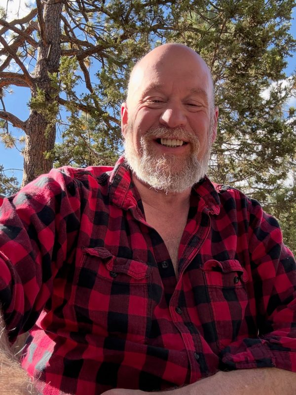 Stephen Ango Oliver smiling outdoors in a red plaid flannel shirt under pine trees